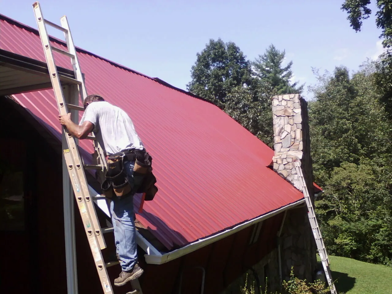 Roofer on a ladder beside a red metal roof and stone chimney — Soffit & Fascia Repair by Roofing Brothers in Kendall Park